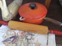 Photo showing orange Le Creuset saucepan with wooden handle, vintage rolling pin with red handles, carved wooden duck, and towel with bird and floral pattern on a wooden table.