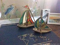 Photo showing two brass and stained glass sailboat figurines of different sizes on a floor with partial wall art in the background