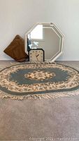 Wool rug with intricate floral and geometric patterns, fringed edges, placed on carpeted floor alongside octagonal mirror and rectangular clock.