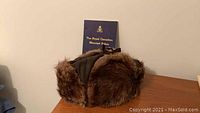 Muskrat fur hat with soft brown fur and fabric lining shown with a navy blue history book standing upright behind it.