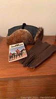Muskrat hat, The Mounties book, and leather gloves displayed on a wooden surface, showing the overall contents of the lot.