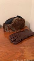 Photo showing the muskrat fur hat and dark brown leather gloves lying on a wooden surface.
