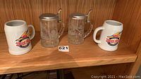Two ceramic beer mugs with Carl's Beer Fest logos and two clear glass beer steins with metal lids on wooden shelf
