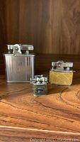Three vintage lighters on wooden surface: largest silver Ronson 'Whirlwind', middle yellow miniature Pacific, smallest Japanese Pygmy mini lighter