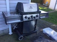 Front view of Broil King gas BBQ grill with extended fold-down side shelves, black and silver finish, and two wheels visible.