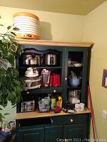 Wide view of black cabinet displaying various kitchen appliances and utensils including crock pots, toaster, blender and cups.
