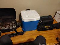 Portable black propane grill with legs and handle beside coolers and rolled camping gear