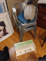 Two stacked silver metal chairs with blue cushioned seats, a hanging globe light fixture placed on top, and a small white wooden hanging shelf with green drawers and hooks on the floor in front of the chairs.