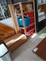 Three wooden shelving unit parts stacked next to complete wood shelving with plastic storage bins