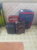 Four suitcases of different colors displayed upright against a mattress. Includes 3 soft shell cases (navy/red, rust red, brown) and 1 black hard shell case.