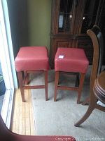 Two wooden stools with red cushioned seats positioned side by side near a sliding glass door, floor visible, minor wear visible on seats and legs.