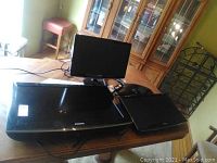 Three Samsung display devices on a wooden table: two monitors and one flat screen TV, all black, cables visible.