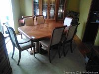 Full view of solid wood rectangular dining table and six chairs arranged around it in a carpeted room, light coming from window nearby
