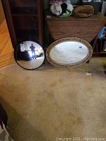 Two mirrors side by side on carpeted floor: a gold gilded oval mirror and a round domed safety mirror.