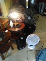 Full view of the brass table lamp on a round wooden side table next to the large black fan, white fan, and black fan heater on the carpeted floor