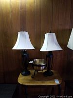 Two black metal table lamps with white shades and a brass-finished metal bowl on black stand on wooden table against wood panel wall