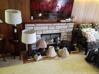 Various lamps and lamp shades grouped together in a living room against a wood paneled wall and stone fireplace. Includes two matching table lamps (white drum shades, dark wood bases), floor lamp, hanging lamp, and multiple lamp shades.
