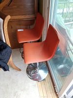 Two orange seat bar stools with chrome metal swivel bases, minor wear and puncture on one seat, placed near glass door with wooden floor.