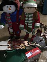 Photo showing two large holiday figures (snowman and girl doll), two metal nutcracker ornaments lying flat, green metal Christmas tree stand with three legs, and plush reindeer and candle set partially visible.