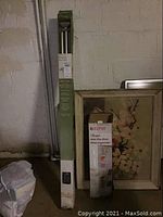 Photo showing boxed Home Edition drapery rod set, framed floral wall art, and boxed Keeper shoe organizer against a concrete wall.