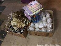 Two boxes seen including champagne bottle shaped candles and a gold-colored tablecloth folded in a box along with scissors and pastel favor boxes.