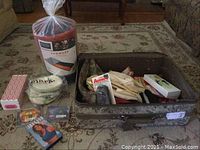 Soft sided suitcase open on carpet with assorted items including boxed items, Avon cosmetic bags, Wenzel Sunward sleeping bag rolled up in packaging, small plastic box, and other accessories.