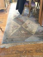 Corner of the area rug showing floral diamond pattern and some furniture legs on hardwood floor.