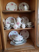 Full view of china set inside cabinet showing various plates, cups, serving pieces with pink floral design and gold trim.