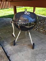 Front view of Weber charcoal kettle grill on tripod legs with lid closed, showing glossy black finish and white handle.