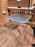 Square glass top patio table with metal base positioned on wooden deck, showing side and top views with scuff marks and some dirt.