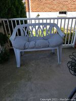 Front view of white plastic outdoor love seat with tufted gray cushion placed on outdoor concrete porch area with railing and brick wall.