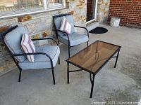 Two blue cushioned metal outdoor chairs and a smoked glass metal frame table on a concrete patio outside a brick house.