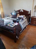 Full view of bed frame with headboard and footboard, mattress, linens, and pillows. Some personal items on bed surface. Dust and wear visible.