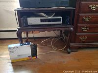 Full view of wood TV stand holding Panasonic VHS recorder with cables underneath and Netgear router box on floor.