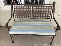 Front view of the metal patio bench with striped cushion placed on the seat, showing lattice backrest and curved armrests.