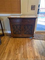 Full view of wooden cabinet with two drawers and two doors with metal scrollwork, set on hardwood floor.
