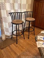 Pair of dark metal spindle backed swivel bar stools with tan round upholstered seats shown standing on wood floor near kitchen area with patterned curtains in background.