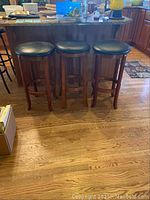Three wooden bar stools with black upholstered seats in a kitchen setting, showing full stools front view and the stools placed next to a kitchen counter.