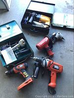 Power tools and toolbox arranged on concrete floor including drills of various types and colors plus a green toolbox with contents