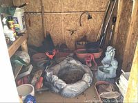 Garden shed corner with a stone-like circular fire pit or planter, child and dog statues, various pots in orange and plastic, and some garden tools against plywood walls.