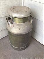 Side view of metal milk can with handles on a concrete porch surface against a beige wall.
