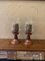 Pair of copper hurricane candle holders on a wooden mantle with a textured background wall.
