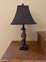 Full view of the table lamp showing the full lamp shade, figurine base, and lamp height on wooden surface near wall.