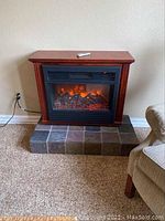Image showing the full electric fireplace with wooden surround and wheels, plugged into a wall outlet, placed on a tiled hearth with carpeted floor.