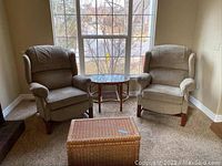 Image of a pair of beige/gray upholstered wingback recliner armchairs with wood bases positioned by a large window, including a wicker chest and small round table.