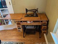 Full front view of the antique Singer sewing machine on wooden table with drawers and cast-iron base. Included is the treadle pedal and the woodworking details.
