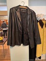Front view of black leather coat hanging on a wall, showing three-button closure and lapels.