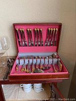 Wooden presentation box open showing silver plated flatware with gold-tone decorative handles lining top and bottom trays