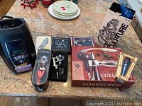 Overall assortment of wine accessories including electric wine chiller, corkscrew in box, pourer in packaging, wine stoppers, and bottle openers displayed on countertop.