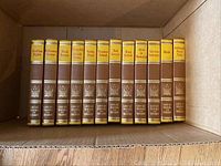 Photo of 12 hardcover books arranged vertically inside a cardboard box, showing the spines with titles and series branding on them.